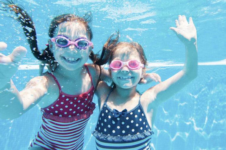 two young girls swimming under water with goggles, waving